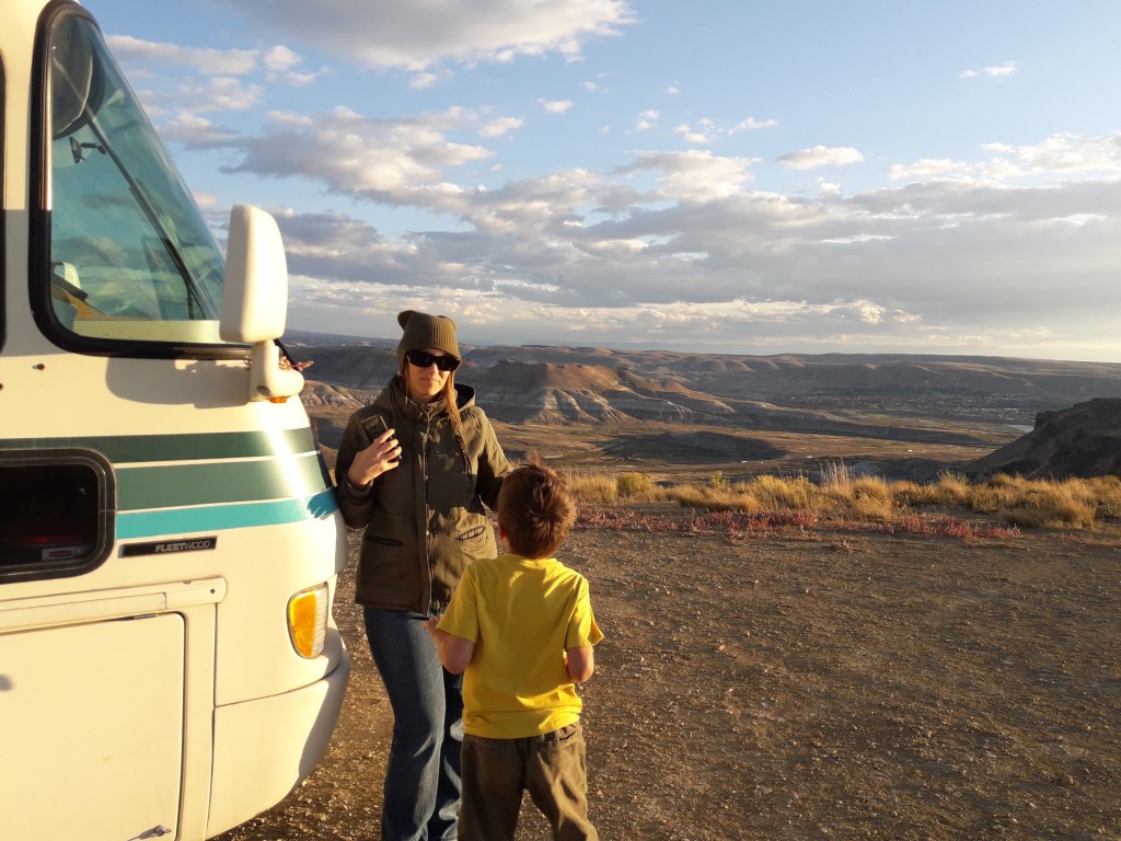 A woman in a green jacket and hat stands next to a recreational vehicle while talking to a young boy in a yellow shirt. They are on a hillside with a scenic view of mountains and valleys under a partly cloudy sky.
