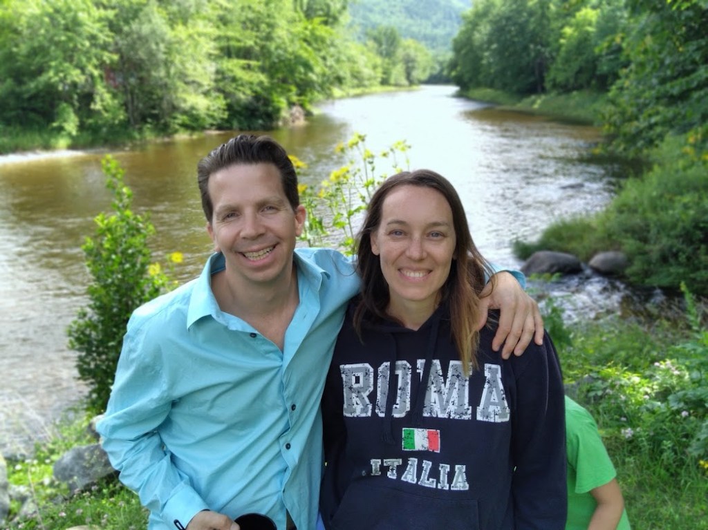A man in a light blue shirt and a woman in a black hoodie pose together by a river surrounded by greenery.