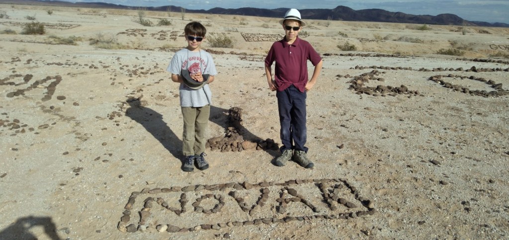 Two boys standing on a sandy area with rock formations spelling out 'NOVAR' on the ground.
