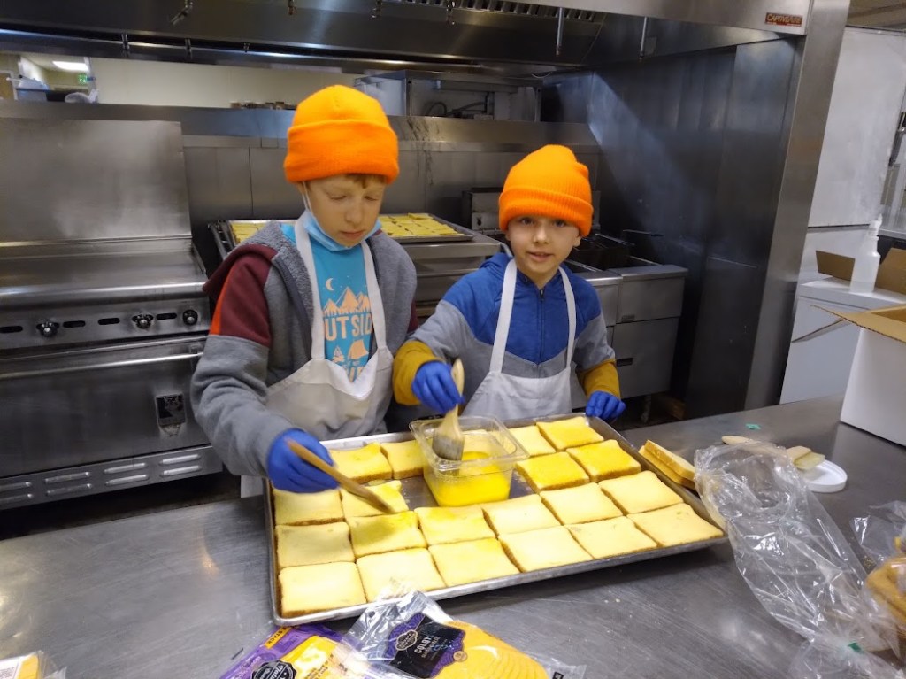 Two children wearing orange beanies and aprons are preparing slices of bread in a kitchen. One child is brushing melted butter onto the bread while the other smiles at the camera. The kitchen has stainless steel appliances and various ingredients visible.