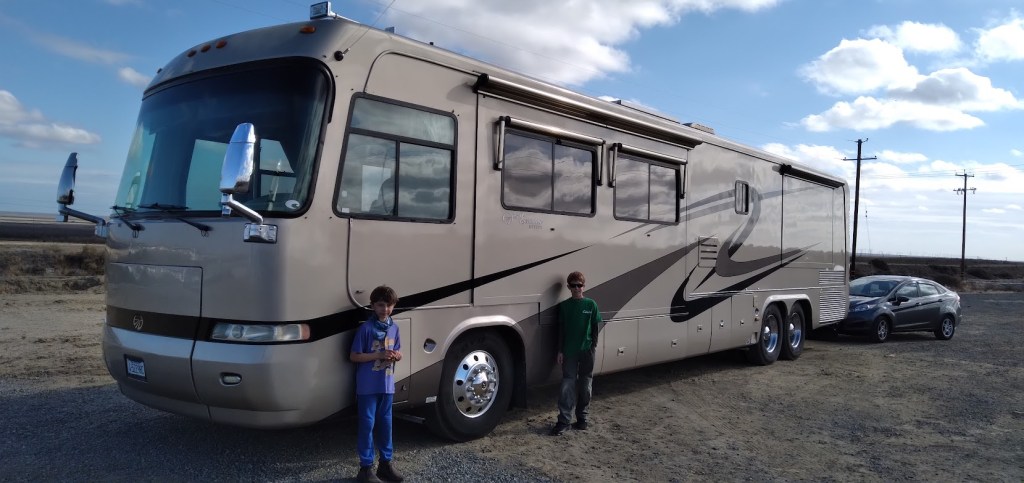A large beige recreational vehicle parked on a gravel surface, with two children standing beside it and a silver car nearby. The sky is partly cloudy.