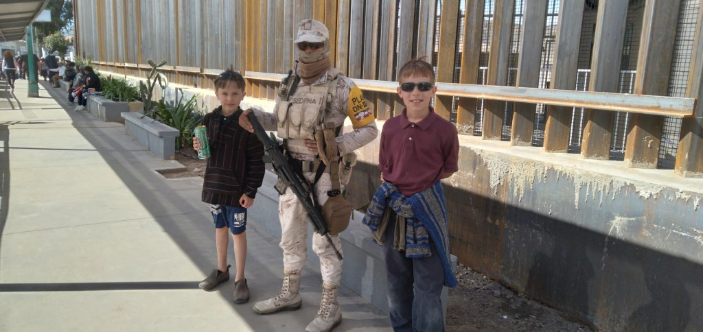 A soldier in military uniform stands between two boys outside a building. One boy is holding a drink while the other wears sunglasses. The setting includes a wall and visitors in the background.