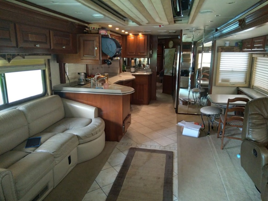 Interior view of a spacious RV with a leather couch, kitchen area, dining table, and decorative lighting.